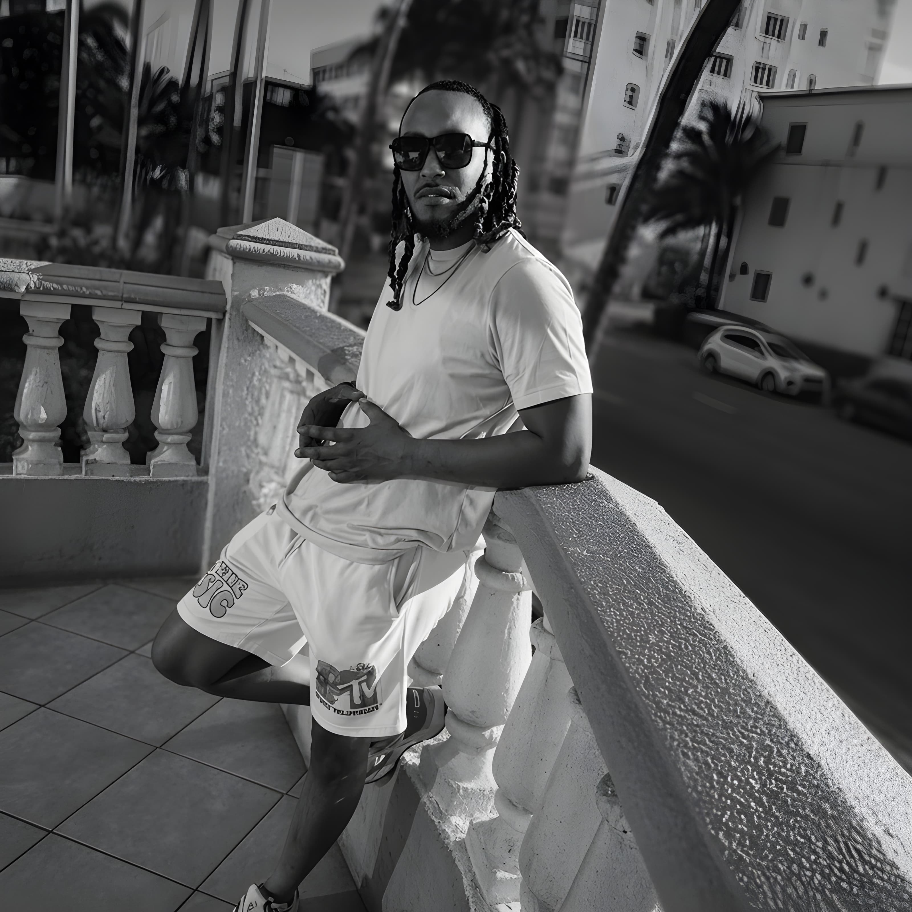 Man with braids and sunglasses leaning on a stone balcony railing in a monochrome shot.