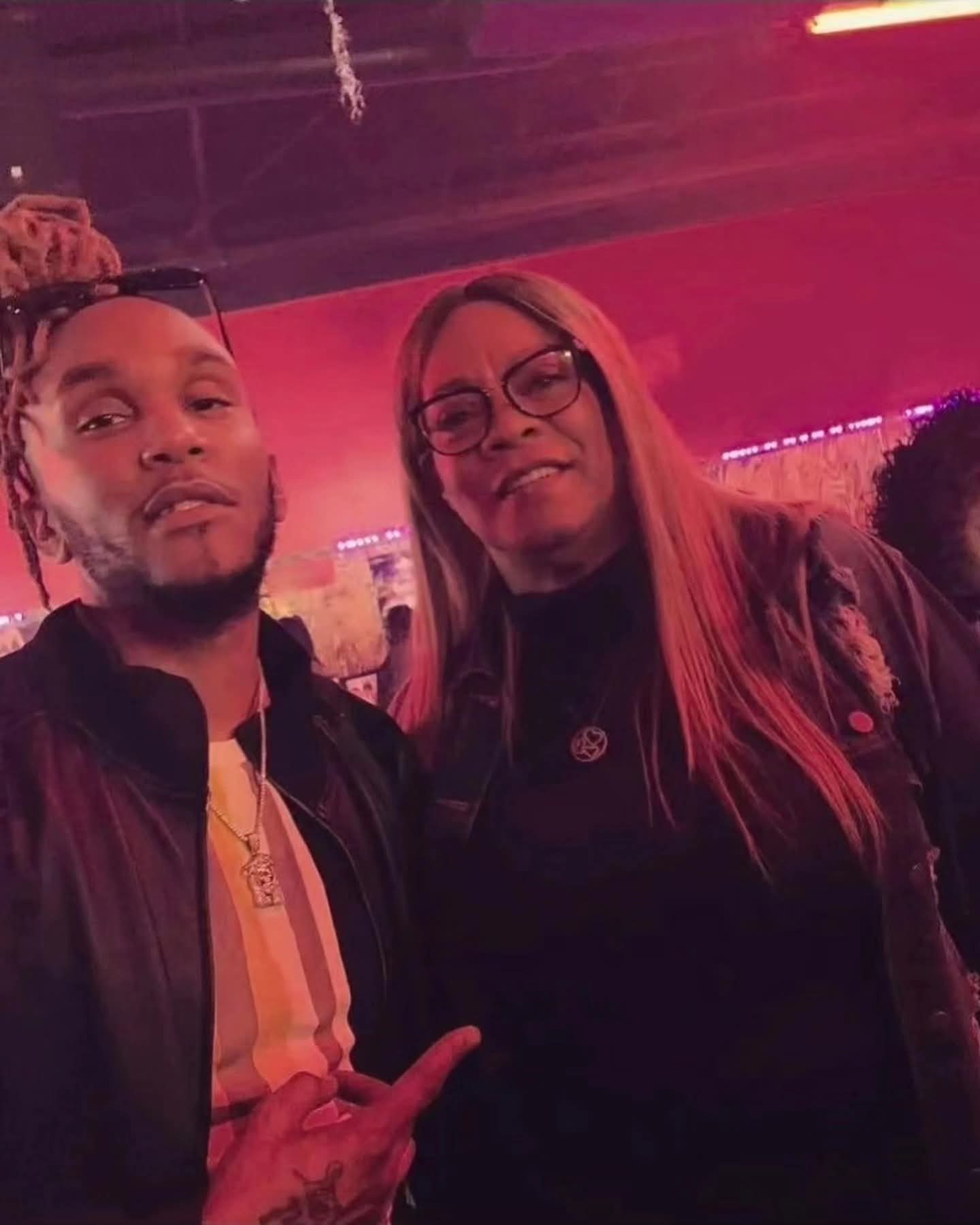 Man with dreadlocks and woman in glasses pose together under vibrant pink indoor lighting.