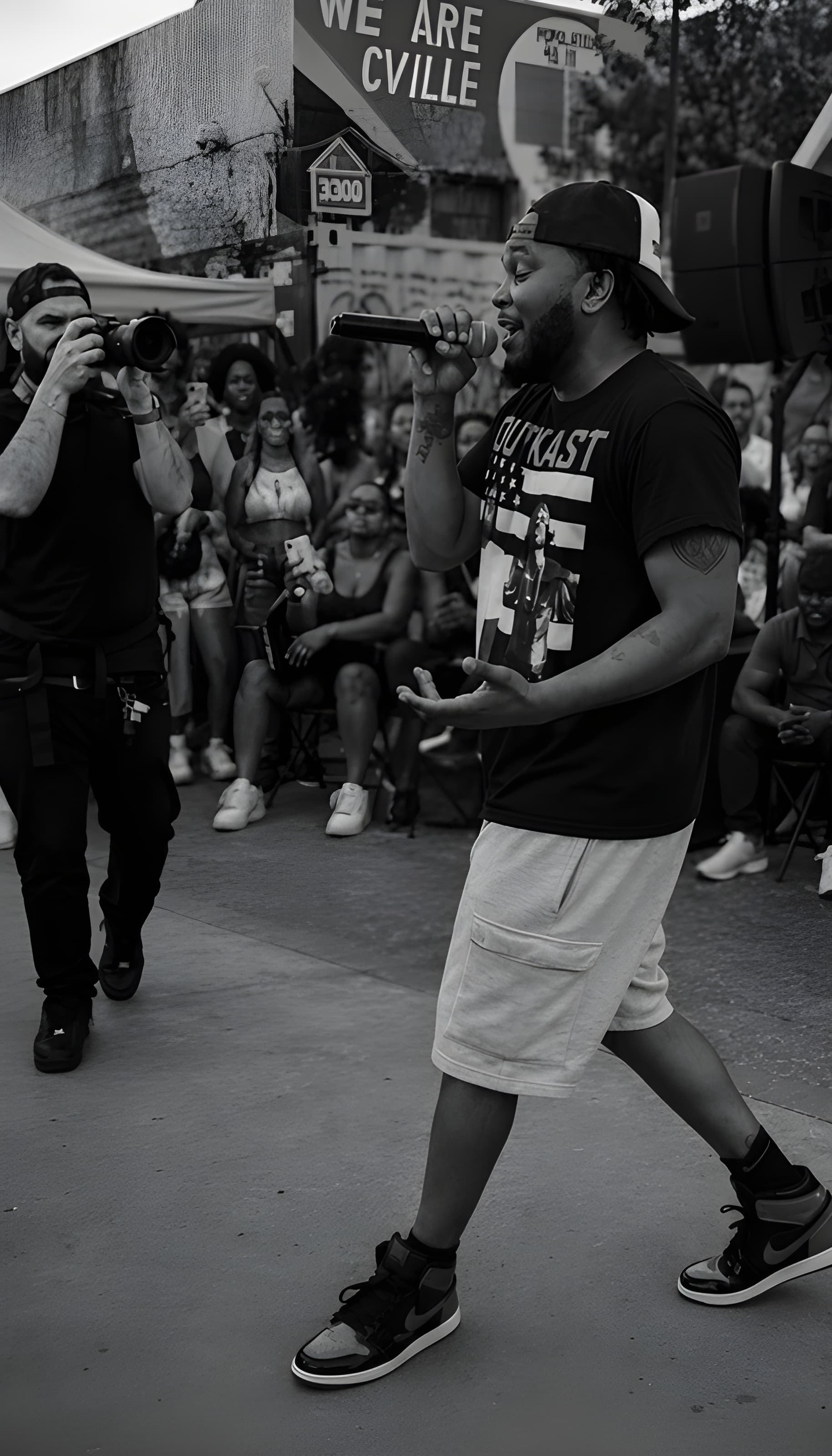 Man in Outkast shirt performs with a microphone outdoors while a photographer captures him.
