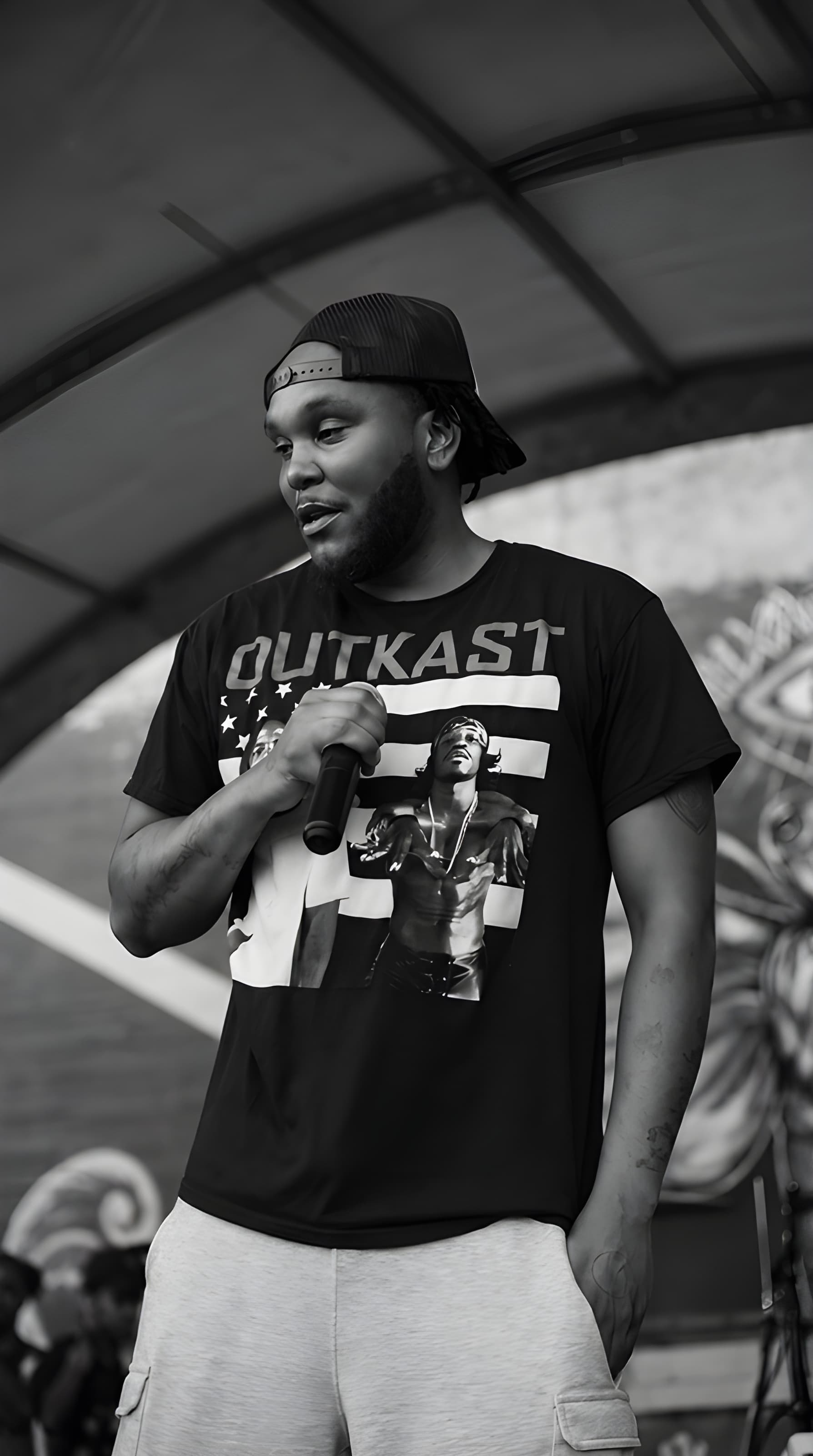 Bearded man in an Outkast t-shirt and backwards cap holds a microphone on stage.