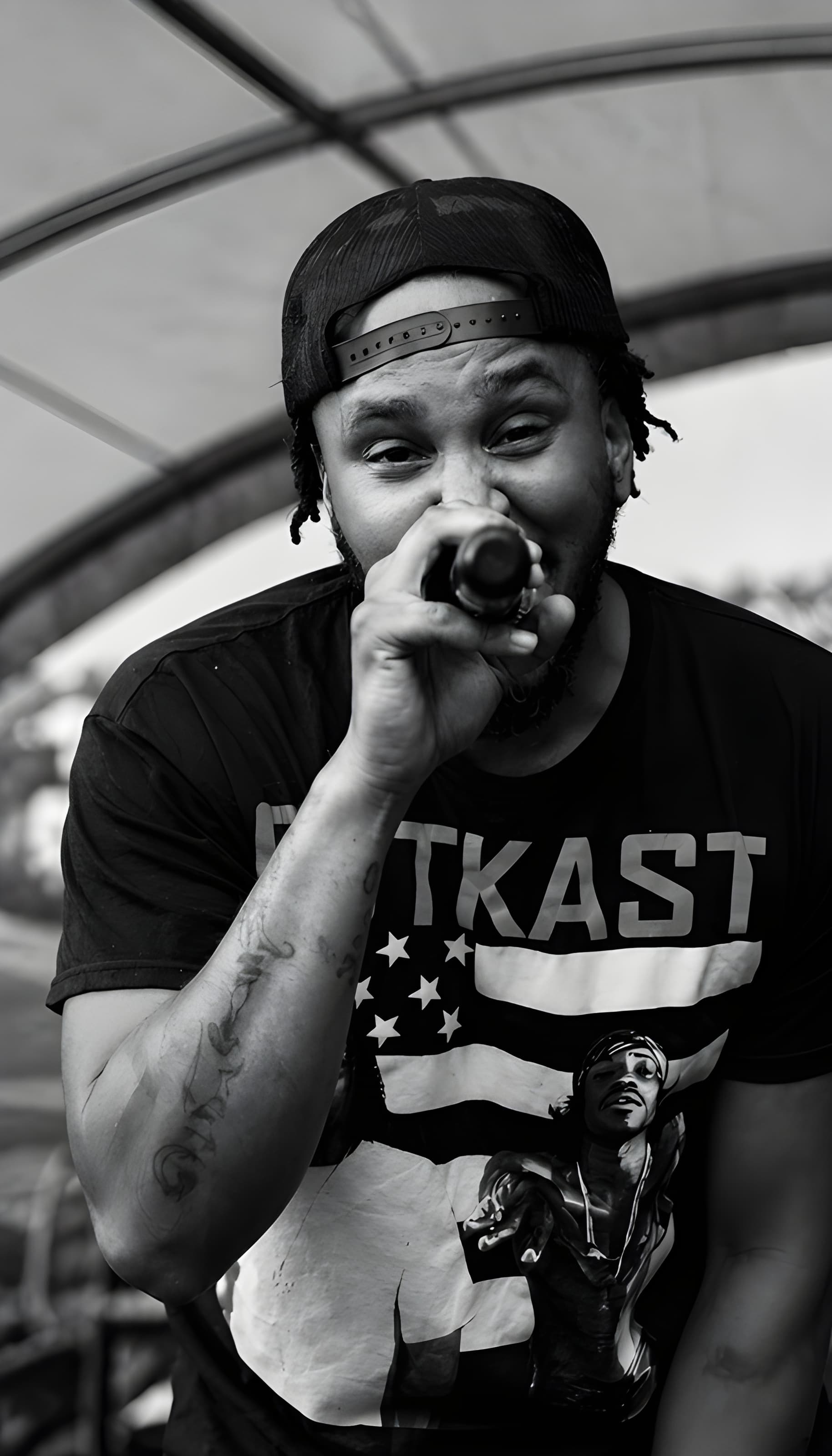 Black and white close-up of a man in an Outkast shirt holding a microphone.